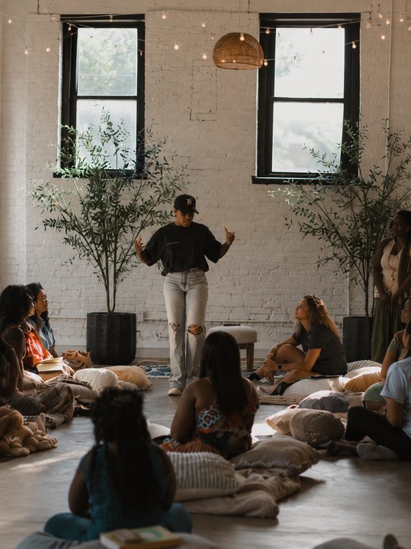 A host speaking to a circle of women seated on the floor of a sunlit loft strung with lights.